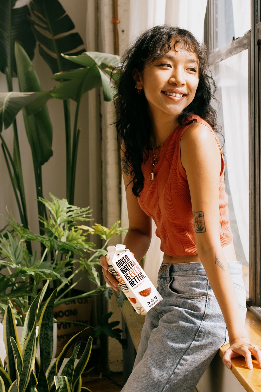 young woman holding Grapefruit Boxed Water in front of a window and houseplants