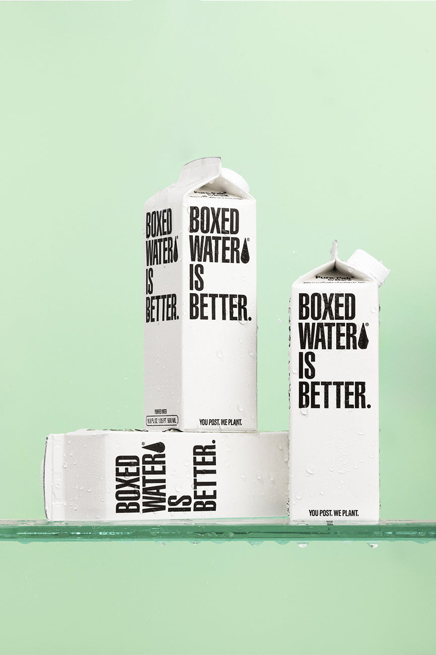 Three boxes of Boxed Water on a glass table in front of a green background 