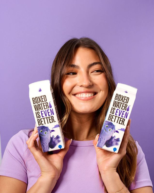 Woman holding 2 IF Boxed Water cartons with purple background
