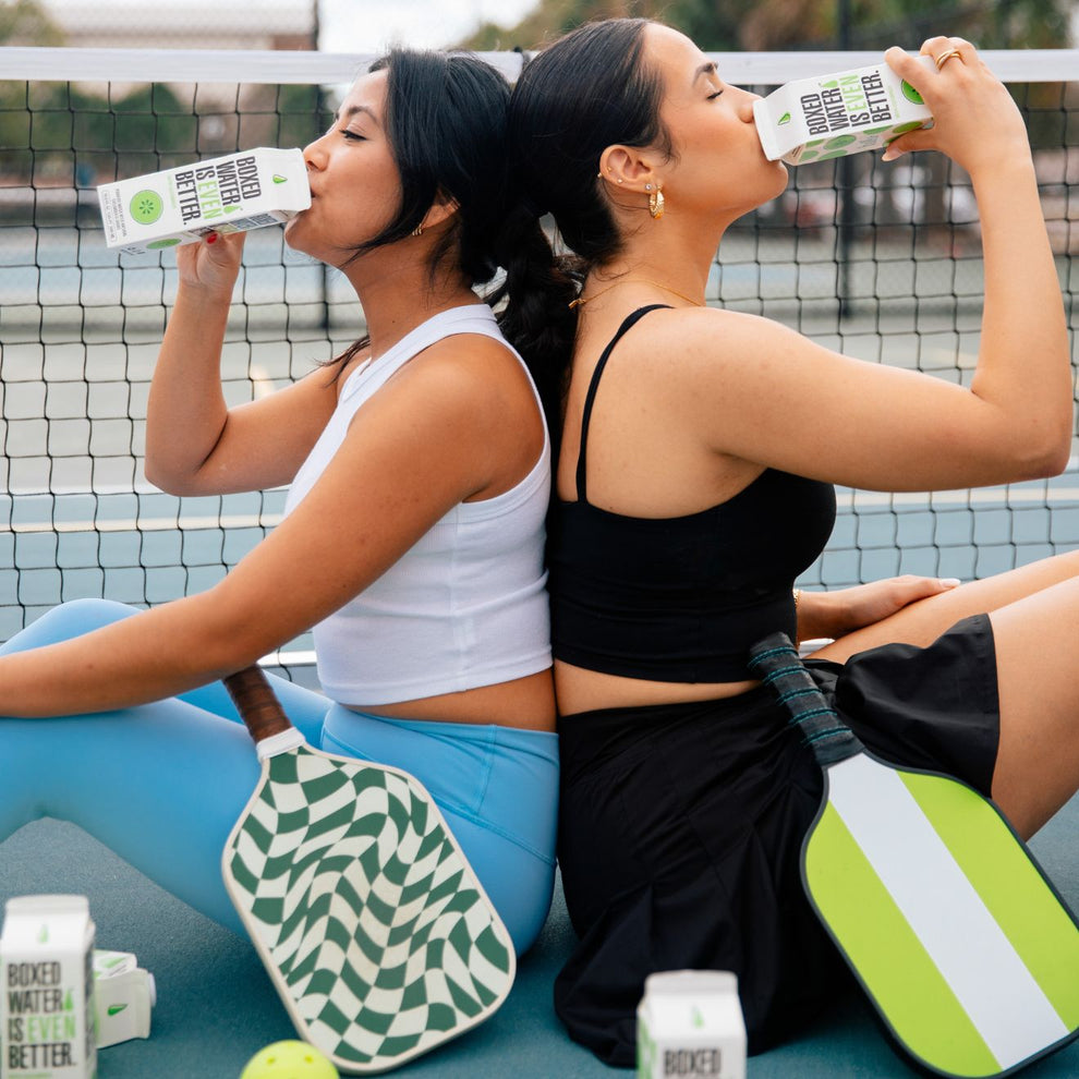 Two woman sitting back to back drinking cucumber Boxed Water on a pickle ball court