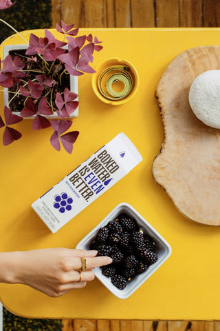 Blackberry flavored boxed water with blackberries in white bowl, flowers on yellow table, charcuterie board on yellow table