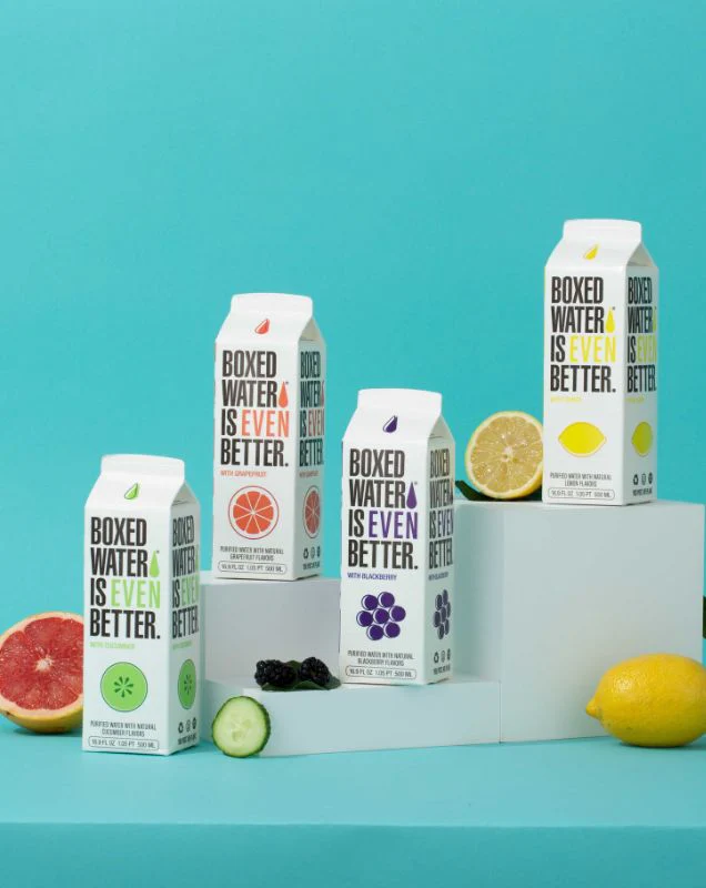 Blackberry, cucumber, grapefruit and lemon flavored boxed waters on pedestal with teal background and fresh fruit displayed