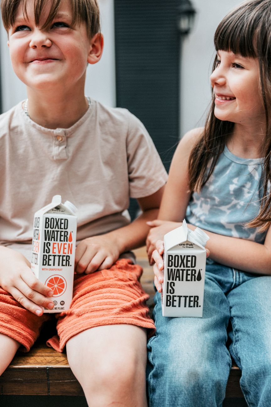 2 yound kids sitting on steps outside holding grapefruit flavored box water and regular boxed water