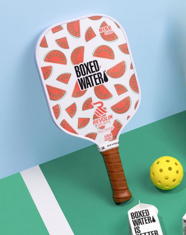 Watermelon pickleball racket and pickleball with boxed water next to them
