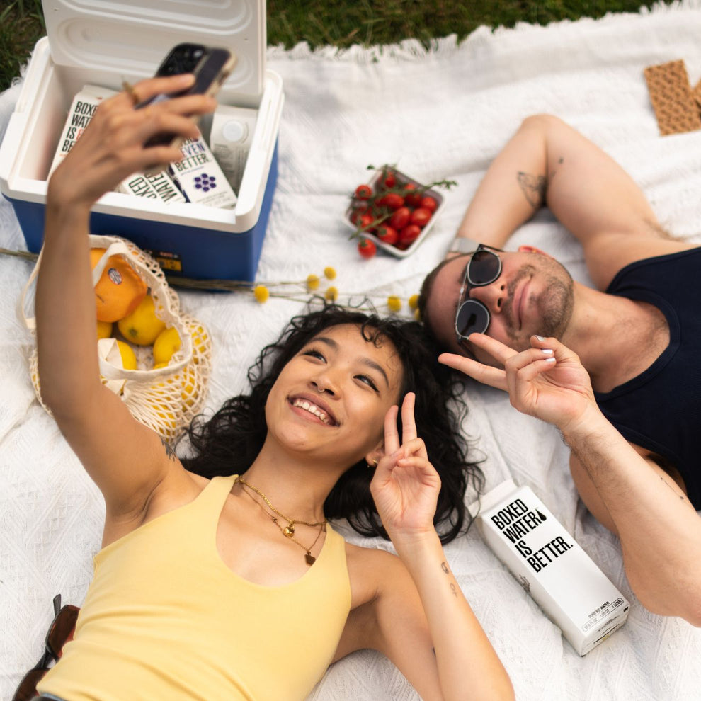 a young man and woman taking a selfie at a picnic with Boxed Water and fruit 