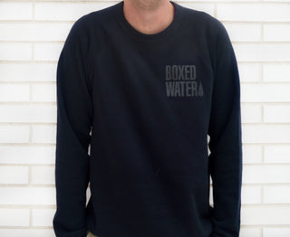 man in black sweatshirt with boxed water logo in front of white brick wall
